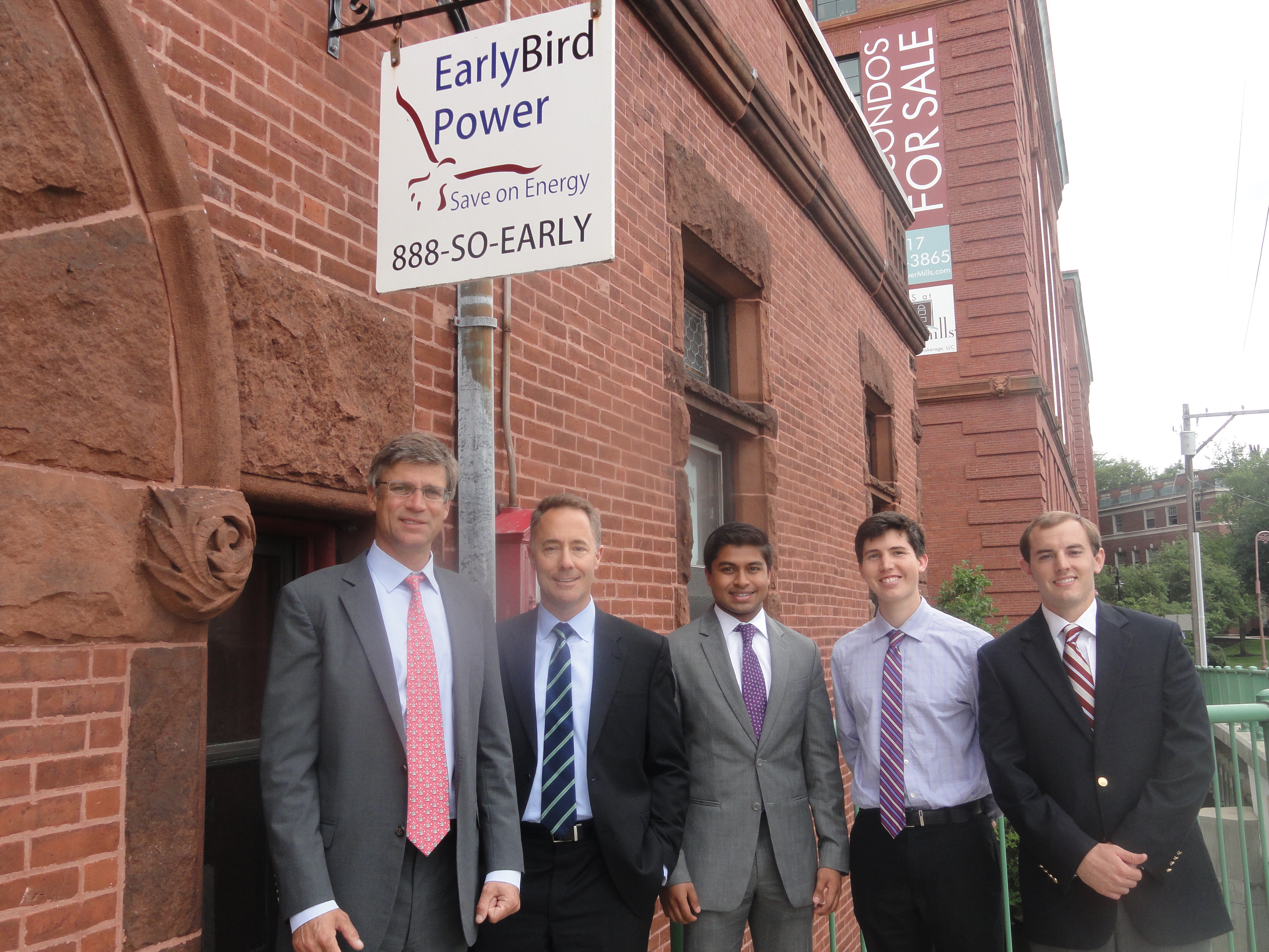 Five men in suits stand outside a brick building with signs about early bird power and a sale.