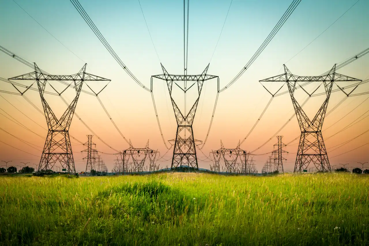 Electric transmission towers stand tall against a colorful sunset sky over a green field.