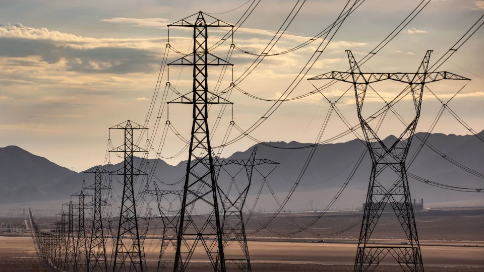 Electricity pylons stand against a dusky sky over mountainous terrain.